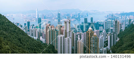 HONG KONG CHINA - DECEMBER 29 2025: Panoramic view of Hong Kong island skyline and Victoria Harbour with skyscrapers and mountains in background on foggy day blue tone HONG KONG CHINA - DECEMBER 29 2025: Panoramic view of Hong Kong island skyline and Victoria Harbour with skyscrapers and mountains in background on foggy day blue tone 140131496