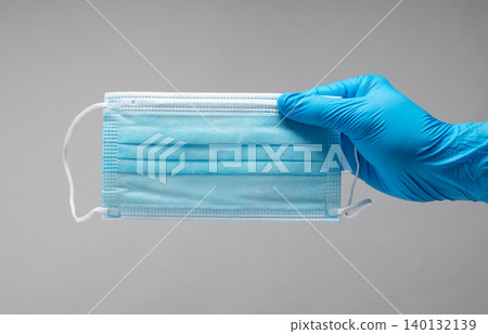 一隻戴著醫用手套的手拿著一次性不織布外科口罩,背景是灰色。 一隻戴著醫用手套的手拿著一次性不織布外科口罩,背景是灰色。 140132139