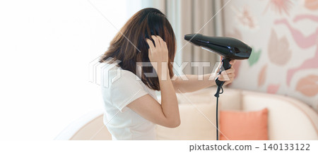 Young woman drying long hair with a hair dryer after a shower in home or hotel room during vacation. Hairstyles and daily morning routine Young woman drying long hair with a hair dryer after a shower in home or hotel room during vacation. Hairstyles and daily morning routine 140133122