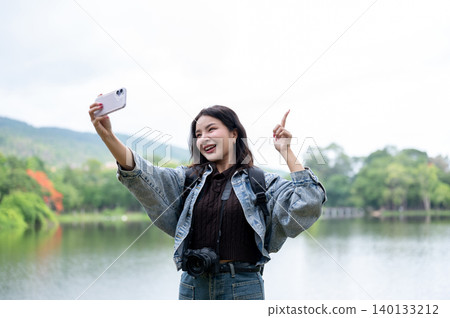 Young asian woman with camera holding phone taking photo pointing up beside reservoir lake in park. Young asian woman with camera holding phone taking photo pointing up beside reservoir lake in park. 140133212