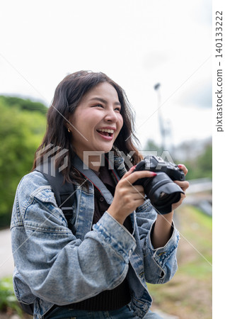 Pretty asian woman in denim holding a camera laughing as looking up while sitting on bench in park. Pretty asian woman in denim holding a camera laughing as looking up while sitting on bench in park. 140133222