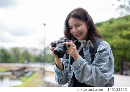 Pretty asian woman in denim holding camera smiling as looking at photos while standing in the park. Pretty asian woman in denim holding camera smiling as looking at photos while standing in the park. 140133223