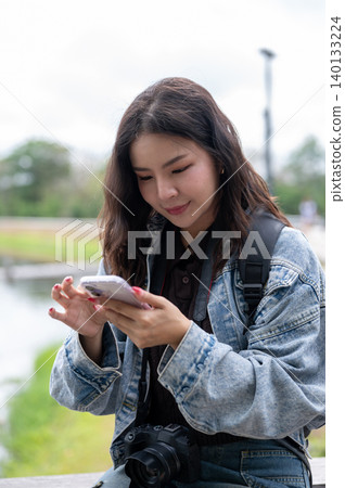 Pretty asian woman looking at smartphone checking photos or chatting while sitting on bench in park. Pretty asian woman looking at smartphone checking photos or chatting while sitting on bench in park. 140133224