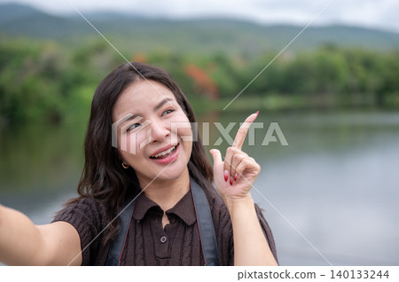 Selfie shot of pretty asian woman holding phone pointing finger up as standing beside lake in park. 140133244