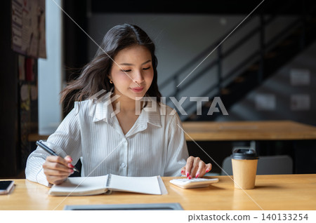 Smart asian woman using calculator holding pen over notebook beside coffee on wooden table in cafe. 140133254