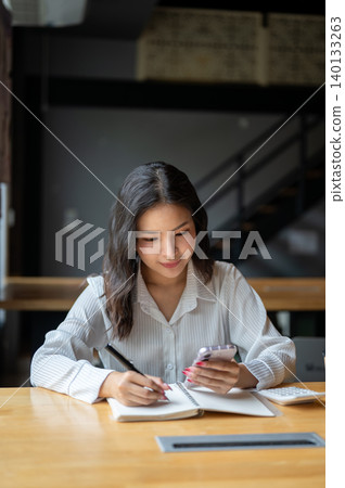 Pretty asian woman looking at phone as writing in notebook beside calculator on wooden table in cafe 140133263