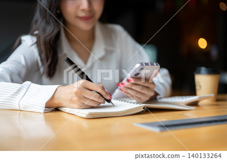 Close up of asian woman looking at phone while writing in notebook beside calculator on wooden table Close up of asian woman looking at phone while writing in notebook beside calculator on wooden table 140133264