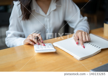 Close up of asian woman looking at phone while writing in notebook beside calculator on wooden table Close up of asian woman looking at phone while writing in notebook beside calculator on wooden table 140133265