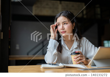 Serious asian woman massaging temple holding pen and coffee beside tablet at wooden table in a cafe. Serious asian woman massaging temple holding pen and coffee beside tablet at wooden table in a cafe. 140133272
