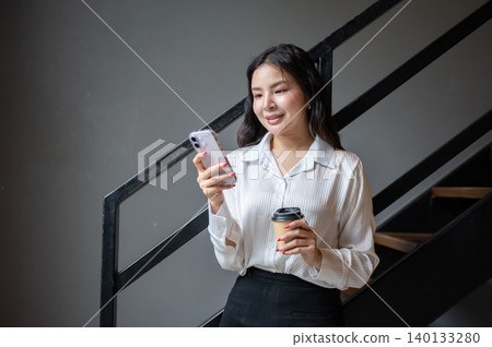Pretty asian woman holding coffee cup looking at phone while standing beside stairs in cafe building Pretty asian woman holding coffee cup looking at phone while standing beside stairs in cafe building 140133280