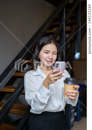 Pretty asian woman holding coffee looking at phone while standing beside the stairs in cafe building Pretty asian woman holding coffee looking at phone while standing beside the stairs in cafe building 140133284