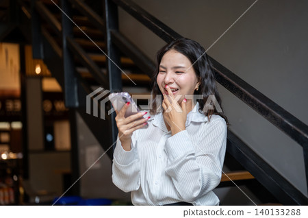 Laughing asian woman covering mouth looking at phone while standing beside stairs in cafe building. Laughing asian woman covering mouth looking at phone while standing beside stairs in cafe building. 140133288