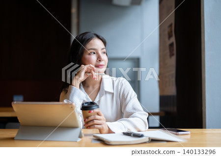Pretty asian woman holding coffee looking away from tablet beside notebook at wooden table in cafe. Pretty asian woman holding coffee looking away from tablet beside notebook at wooden table in cafe. 140133296