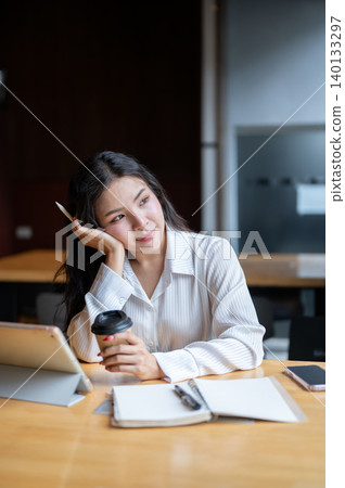 Pretty asian woman holding coffee resting head on hand beside tablet sitting at wooden table in cafe Pretty asian woman holding coffee resting head on hand beside tablet sitting at wooden table in cafe 140133297