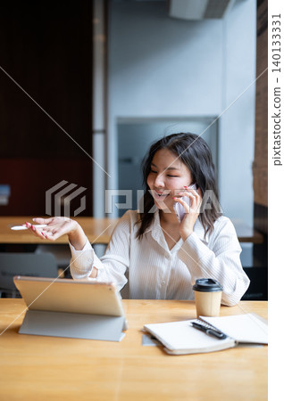 Happy asian woman talking on phone looking at tablet beside coffee sitting at wooden table in cafe. Happy asian woman talking on phone looking at tablet beside coffee sitting at wooden table in cafe. 140133331