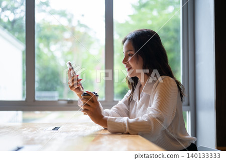 Side view of pretty asian woman looking at phone holding coffee sitting at table beside window in cafe. 140133333