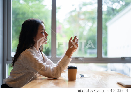 Side view of pretty asian woman watching or playing on phone sitting at table beside window in cafe. 140133334