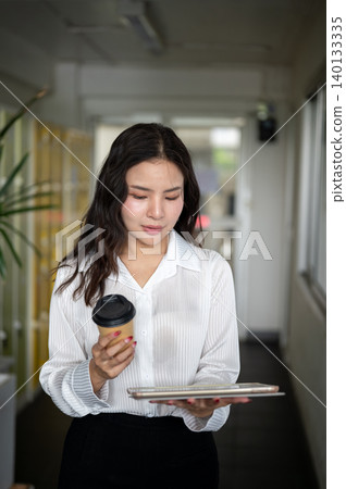 Smart asian woman holding coffee looking at tablet while standing walking in corridor of a building. Smart asian woman holding coffee looking at tablet while standing walking in corridor of a building. 140133335