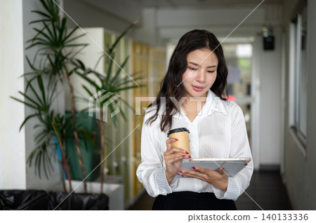 Pretty asian woman holding coffee using a tablet while standing or walking in corridor of building. 140133336