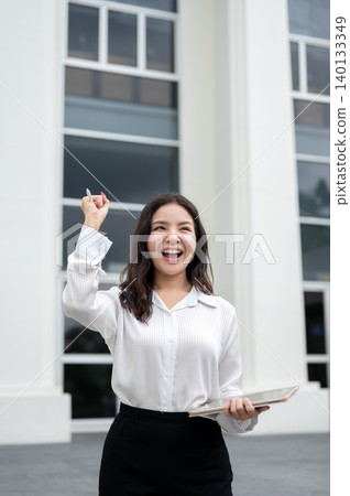 Pretty asian businesswoman holding tablet raising a fist while standing in front of office building. Pretty asian businesswoman holding tablet raising a fist while standing in front of office building. 140133349