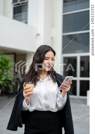 Pretty asian businesswoman holding coffee cup looking at phone while standing outside the building. 140133352