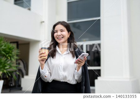 Happy asian businesswoman holding a coffee cup and smartphone while standing outside the building. Happy asian businesswoman holding a coffee cup and smartphone while standing outside the building. 140133355