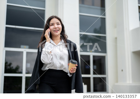 Pretty asian businesswoman holding coffee cup and talking on phone while walking out of the building 140133357