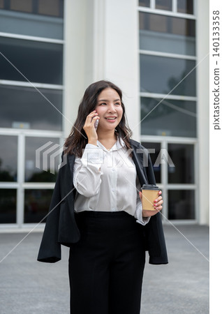 Pretty asian businesswoman holding a coffee cup talking on phone while standing outside the building 140133358