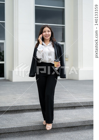 Full body of asian businesswoman holding coffee talking on phone and walking down building's stairs. Full body of asian businesswoman holding coffee talking on phone and walking down building's stairs. 140133359