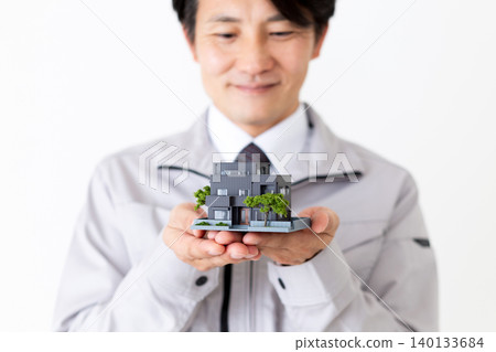 A middle-aged businessman in gray work clothes smiles while holding a model of a house against a white background. A middle-aged businessman in gray work clothes smiles while holding a model of a house against a white background. 140133684