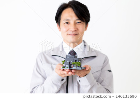 一位身穿灰色工作服的中年商人面帶微笑，手持房屋模型，背景為白色。 140133690