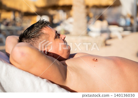 Caucasian man relaxes on a beach lounge chair at a tropical resort under warm sunlight with calm and peaceful expression. Beach vacation and relaxation lifestyle Caucasian man relaxes on a beach lounge chair at a tropical resort under warm sunlight with calm and peaceful expression. Beach vacation and relaxation lifestyle 140136030