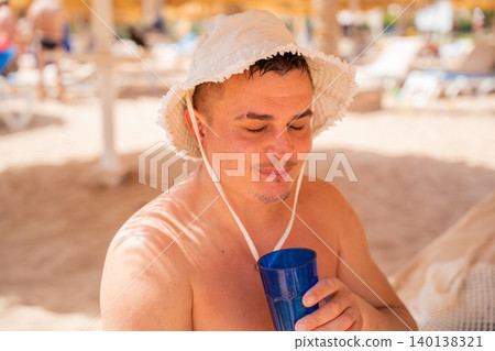 Caucasian man drinks from a blue cup while relaxing at a beach resort in Sharm El Sheikh Egypt with calm and satisfied expression. Beach vacation and relaxation lifestyle Caucasian man drinks from a blue cup while relaxing at a beach resort in Sharm El Sheikh Egypt with calm and satisfied expression. Beach vacation and relaxation lifestyle 140138321