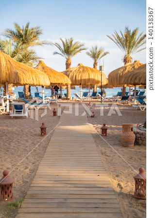 Wooden pathway leads through a beach resort in Sharm El Sheikh Egypt with palm trees straw umbrellas and sandy coastline. Travel and resort lifestyle Wooden pathway leads through a beach resort in Sharm El Sheikh Egypt with palm trees straw umbrellas and sandy coastline. Travel and resort lifestyle 140138367