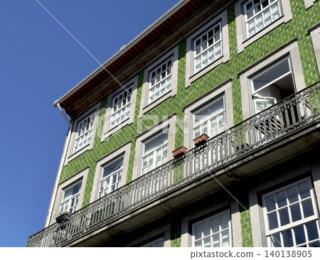 Corner view of green azulejo building with balconies under blue sky. Traditional Portuguese facade displays patterned tiles and window details. Corner view of green azulejo building with balconies under blue sky. Traditional Portuguese facade displays patterned tiles and window details. 140138905