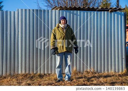 Man wearing warm coat and beanie listening to music with headphones on farm. Rural lifestyle in autumn. Farmer standing by metal fence, relaxing and enjoying sound outdoors. Man wearing warm coat and beanie listening to music with headphones on farm. Rural lifestyle in autumn. Farmer standing by metal fence, relaxing and enjoying sound outdoors. 140139863