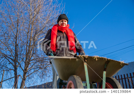 Gardener man pushing wheelbarrow in garden during autumn. Caucasian man working outdoors on sunny day. Seasonal yard maintenance at home. Agricultural hobby and manual labor concept. Gardener man pushing wheelbarrow in garden during autumn. Caucasian man working outdoors on sunny day. Seasonal yard maintenance at home. Agricultural hobby and manual labor concept. 140139864