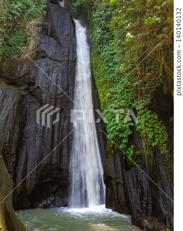 Tall single stream waterfall dropping down dark volcanic rock cliff covered with green vines in tropical jungle Tall single stream waterfall dropping down dark volcanic rock cliff covered with green vines in tropical jungle 140140132