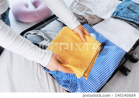 a woman handles travel preparation for a summer vacation, packing a bright yellow towel into an open suitcase filled with clothes and tourism essentials a woman handles travel preparation for a summer vacation, packing a bright yellow towel into an open suitcase filled with clothes and tourism essentials 140140884