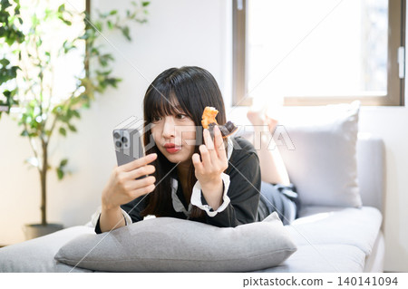 Japanese woman eating a donut while looking at her smartphone in bed Japanese woman eating a donut while looking at her smartphone in bed 140141094