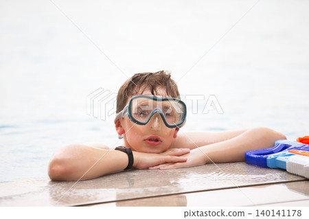 Young boy relaxes by the pool wearing goggles during summer at a recreational center with water toys nearby Young boy relaxes by the pool wearing goggles during summer at a recreational center with water toys nearby 140141178