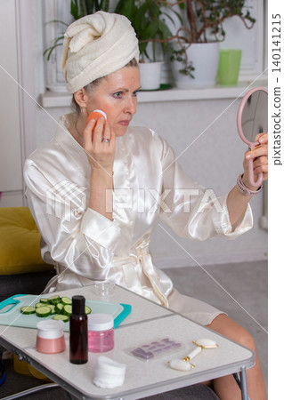 Woman applying skincare products in a bright room while sitting at a table with cucumber slices nearby Woman applying skincare products in a bright room while sitting at a table with cucumber slices nearby 140141215