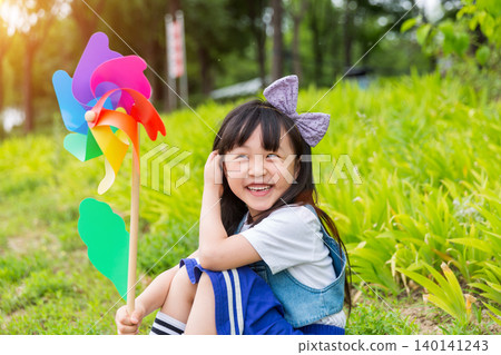Happy Young East Asian Girl with Purple Bow Sitting in Sunny Green Meadow Holding Colorful Rainbow Pinwheel with Joyful Smile and Bright Summer Vibes. Happy Young East Asian Girl with Purple Bow Sitting in Sunny Green Meadow Holding Colorful Rainbow Pinwheel with Joyful Smile and Bright Summer Vibes. 140141243