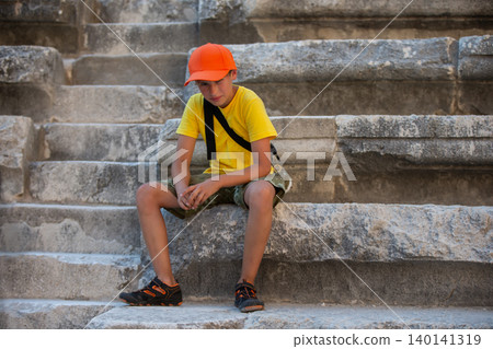 Young boy in bright clothes resting on stone steps of ancient ruins during daytime exploration Young boy in bright clothes resting on stone steps of ancient ruins during daytime exploration 140141319