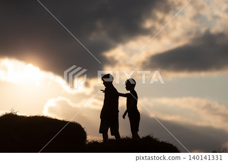 Two boys play on hay bales at sunset with a dramatic sky in the background, creating a serene rural atmosphere Two boys play on hay bales at sunset with a dramatic sky in the background, creating a serene rural atmosphere 140141331
