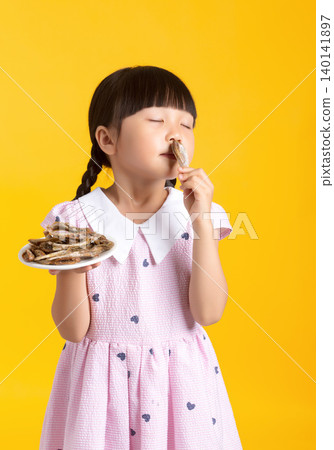 Cute Asian little girl eating dried fish snack, studio yellow background, happy child enjoying food, childhood nutrition concept 140141897