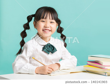 Asian little schoolgirl writing in notebook at desk, focused child studying with books on blue background Asian little schoolgirl writing in notebook at desk, focused child studying with books on blue background 140141903