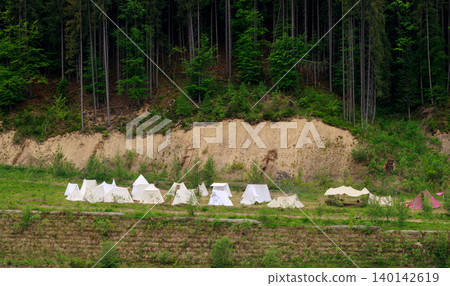 historical reenactment village reconstruction festival camp tent featuring numerous canvas tents arranged in a grassy clearing beside a steep, wooded hillside 140142619
