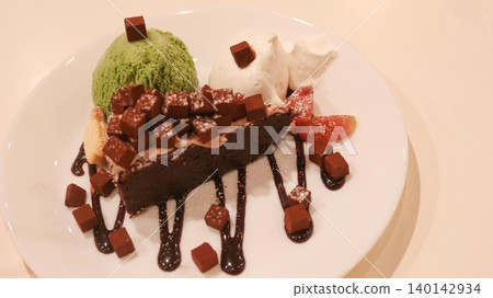 close up of a chocolate brownie cake with green tea matcha ice cream and cream on the sides. Melt chocolate suace and chocolate dice added as decoration on a white plate with copy space close up of a chocolate brownie cake with green tea matcha ice cream and cream on the sides. Melt chocolate suace and chocolate dice added as decoration on a white plate with copy space 140142934
