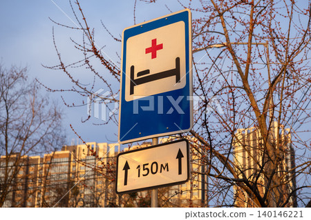 Hospital road sign with a red cross and bed symbol indicates medical services nearby with a distance marker of one hundred fifty meters in a city environment under a clear blue sky Hospital road sign with a red cross and bed symbol indicates medical services nearby with a distance marker of one hundred fifty meters in a city environment under a clear blue sky 140146221